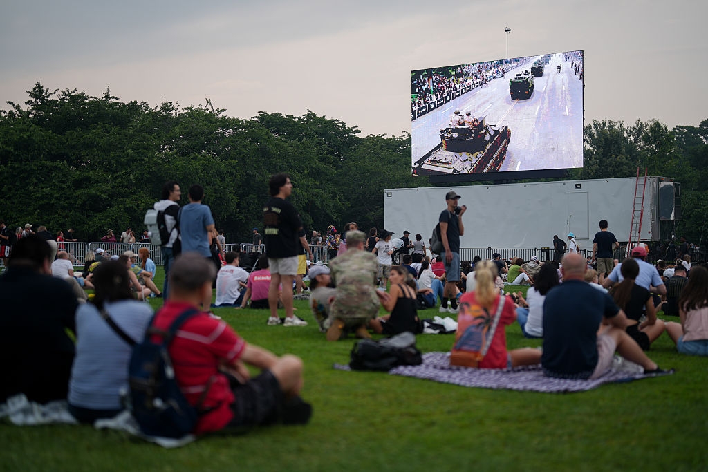 People sitting on grass watching a large screen showing a parade with military vehicles