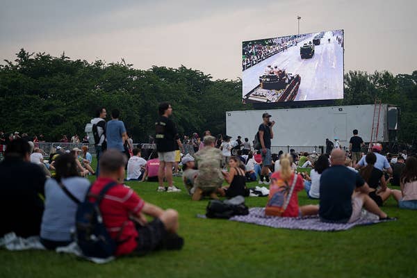 People sitting on grass watching a large screen showing a parade with military vehicles