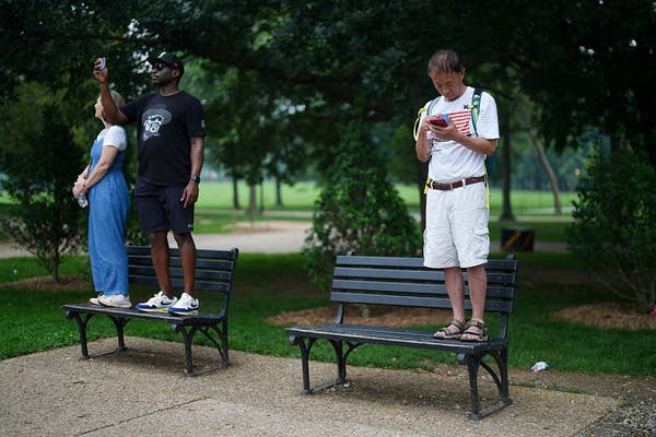 Three people stand on park benches focused on their phones, seemingly unaware of their surroundings
