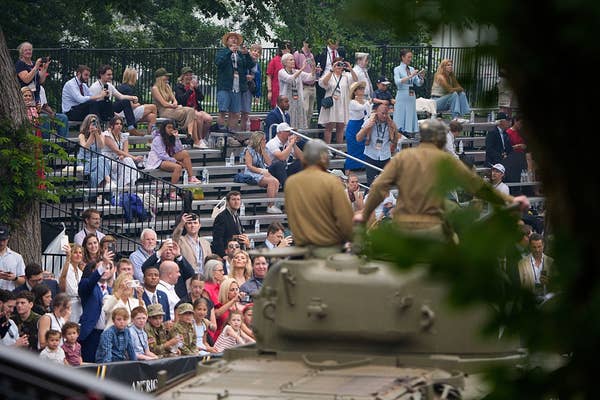 People gather in a park, seated in bleachers, watching a historical military vehicle with two people standing on it