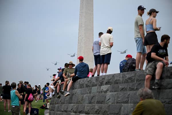 People gathered on a stone structure near the Washington Monument, watching helicopters fly by in the sky
