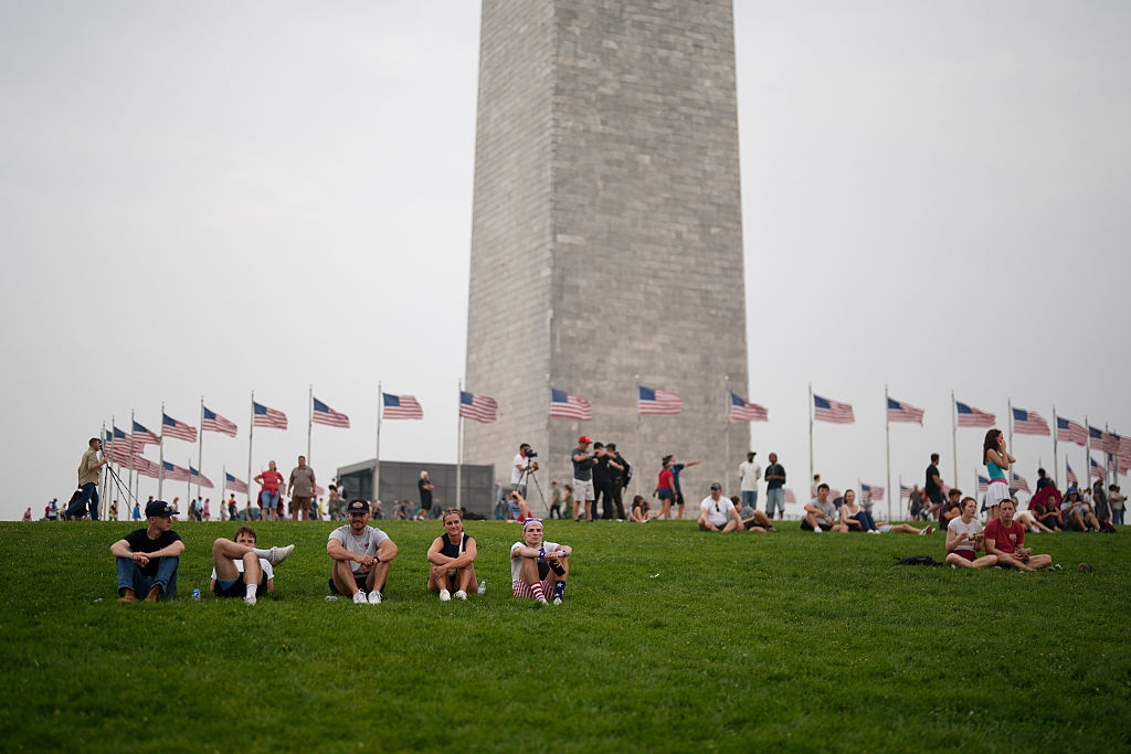 People sit on a grassy area in front of the Washington Monument, surrounded by U.S. flags