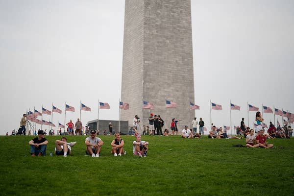 People sit on a grassy area in front of the Washington Monument, surrounded by U.S. flags