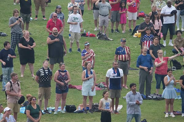 A group of people stands outside with hands over hearts, participating in a patriotic event. Some wear flag-themed attire