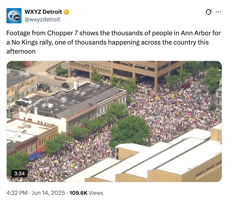 Aerial view showing a large crowd rallying in Ann Arbor for a "No Kings" protest. Dense gathering fills streets, seen from above
