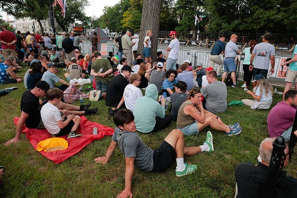 A group of people sit and stand on grass at an outdoor event, casually dressed, with trees and a crowd barrier in the background