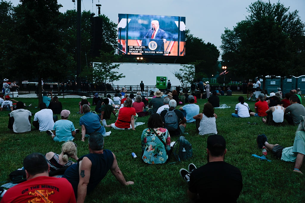 People sitting on grass watching a large outdoor screen displaying a speaker at an event. Trees and equipment are visible in the background