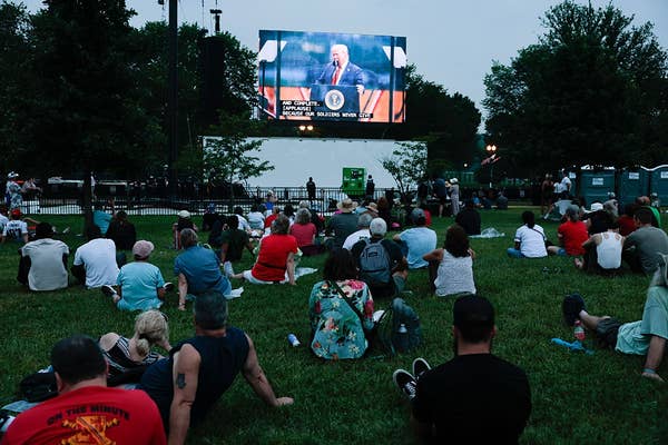 People sitting on grass watching a large outdoor screen displaying a speaker at an event. Trees and equipment are visible in the background