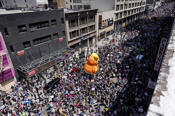 Aerial view of a large protest with a giant inflatable duck in the crowd, filling a city street with people holding signs