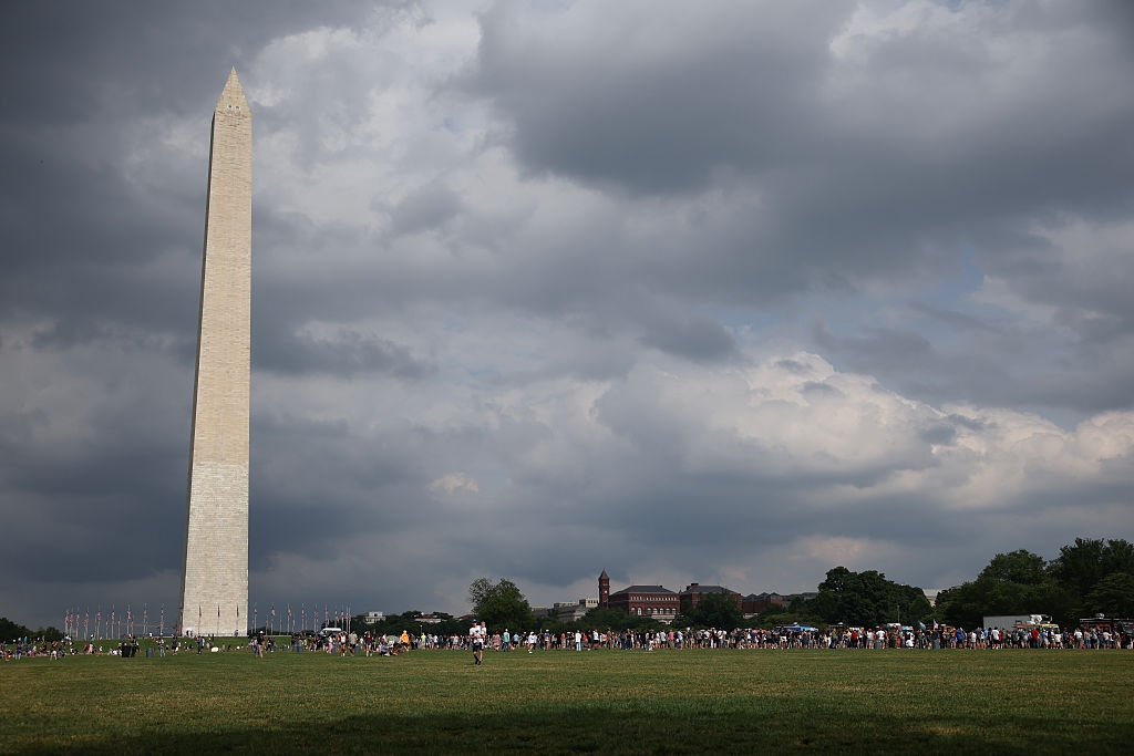 The Washington Monument stands tall against a cloudy sky, with crowds of people gathered on the grass below