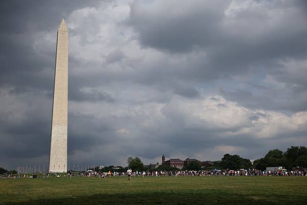 The Washington Monument stands tall against a cloudy sky, with crowds of people gathered on the grass below
