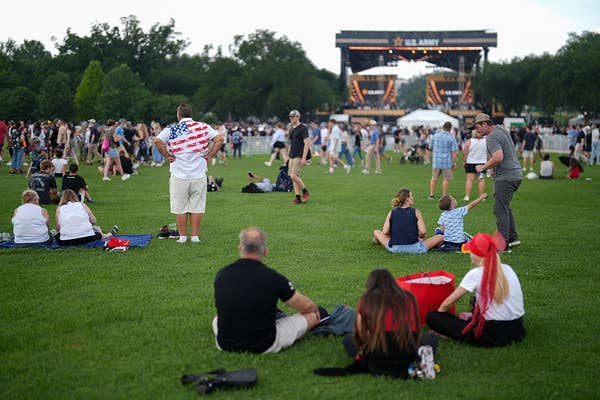 People relaxing on a grassy field at an outdoor concert with a stage visible in the background
