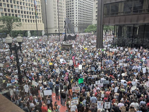 Large crowd gathered in a city square for a protest, holding various signs and banners, surrounded by tall buildings