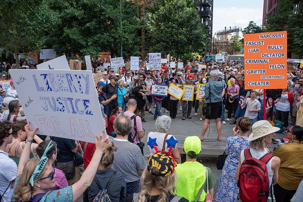 A large crowd gathered at a protest, holding various signs with messages advocating for justice and criticizing a public figure