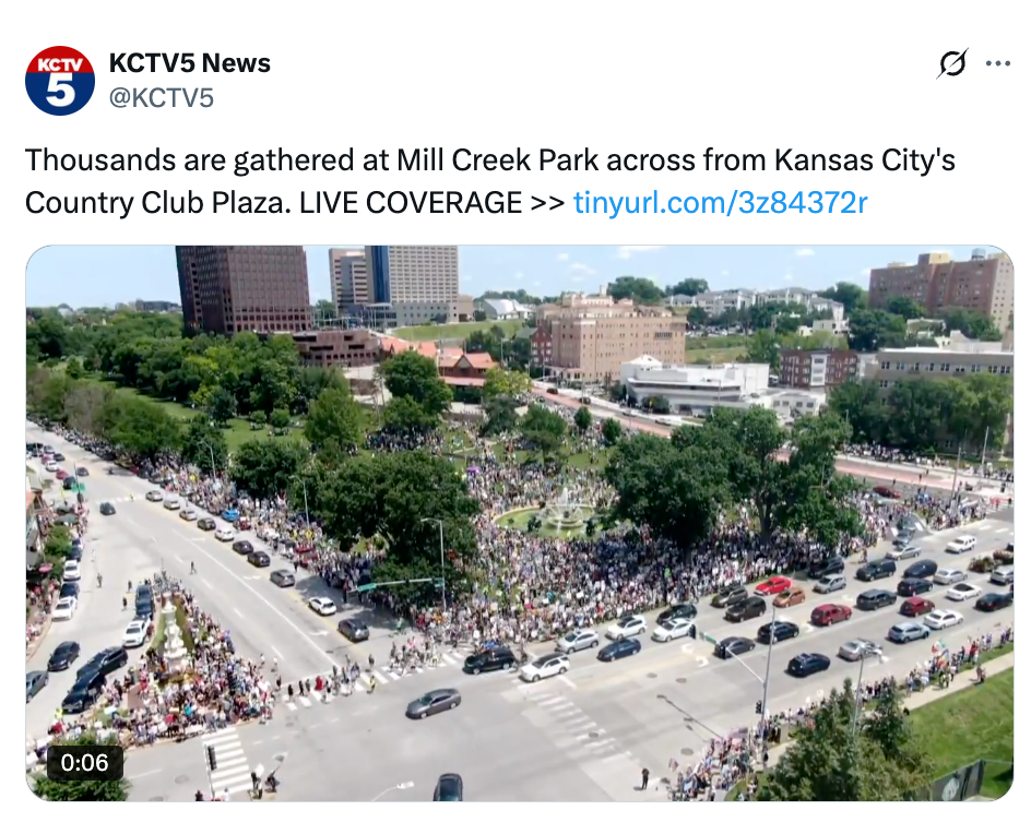 Aerial view of a large crowd gathered at Mill Creek Park, Kansas City, for a news event. Buildings and roads surround the park
