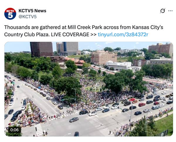 Aerial view of a large crowd gathered at Mill Creek Park, Kansas City, for a news event. Buildings and roads surround the park