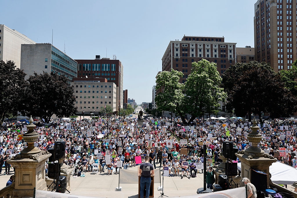 Large crowd gathered for a public protest or rally in an urban square with many holding signs, viewed from behind a speaker at a podium