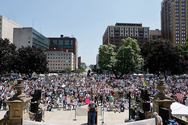 Large crowd gathered for a public protest or rally in an urban square with many holding signs, viewed from behind a speaker at a podium