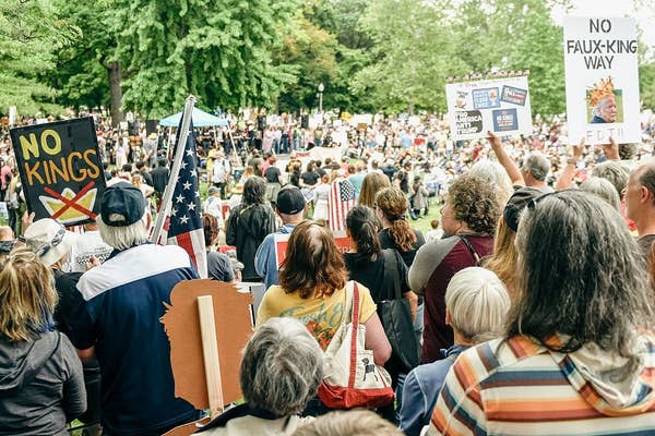 A large crowd gathers at an outdoor protest, holding signs with various messages, including &quot;No Kings&quot; and &quot;No Faux-King Way.&quot;