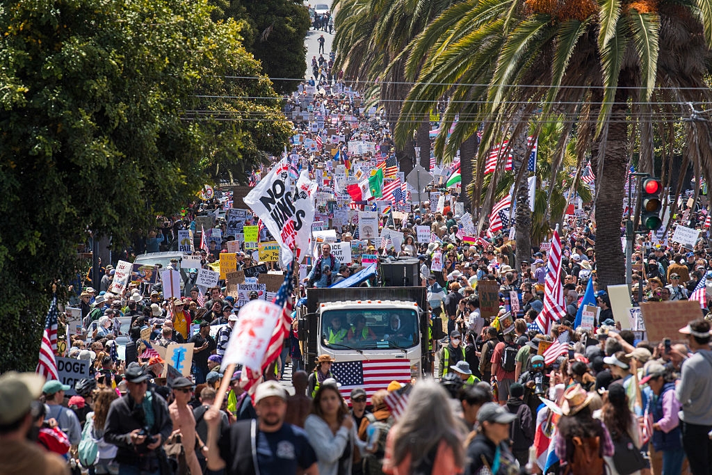 A large crowd of people march in protest on a city street, holding signs and flags