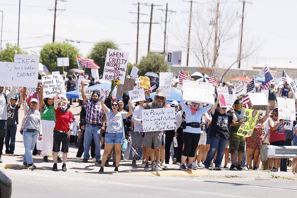 A large group of people hold various protest signs during a demonstration on a street. Some participants wave American flags