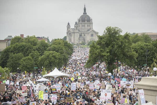 Large crowd gathers for a protest with signs in a city park, historic building in the background