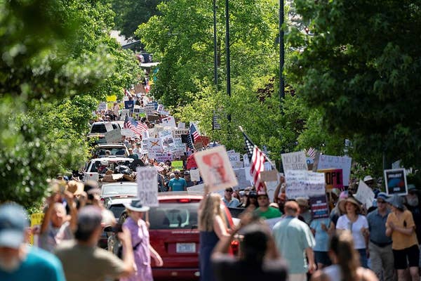 A large crowd of protesters holding various signs and flags marches down a street surrounded by trees