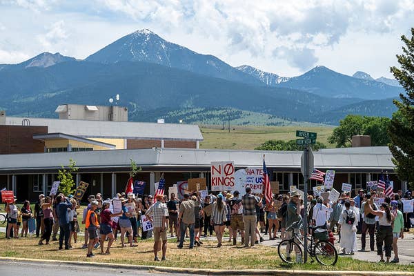 A crowd holds signs at an outdoor protest near a building with mountains in the background. Signs include messages like &quot;No Kings&quot; and &quot;Hate Has No Home Here.&quot;