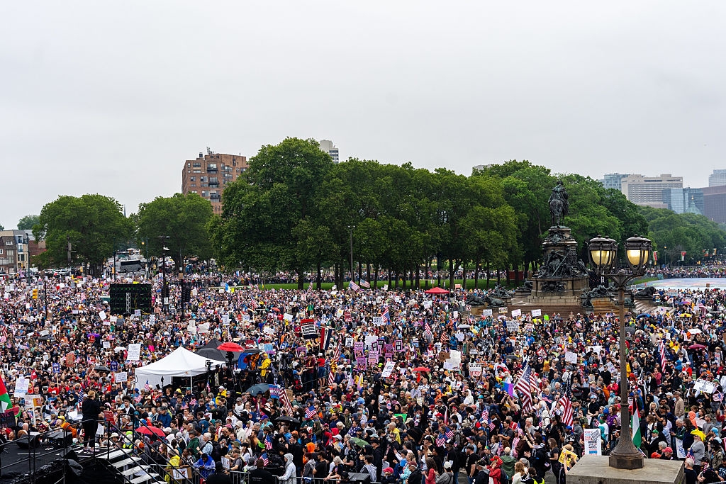 Large crowd gathered at an outdoor event, holding signs and flags, near a statue and trees, with buildings visible in the background