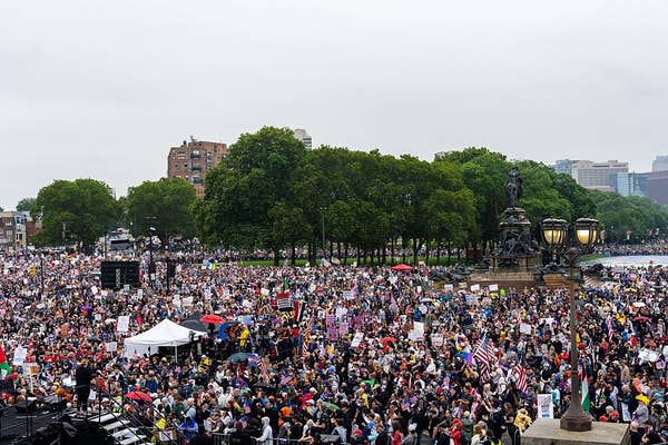 Large crowd gathered at an outdoor event, holding signs and flags, near a statue and trees, with buildings visible in the background