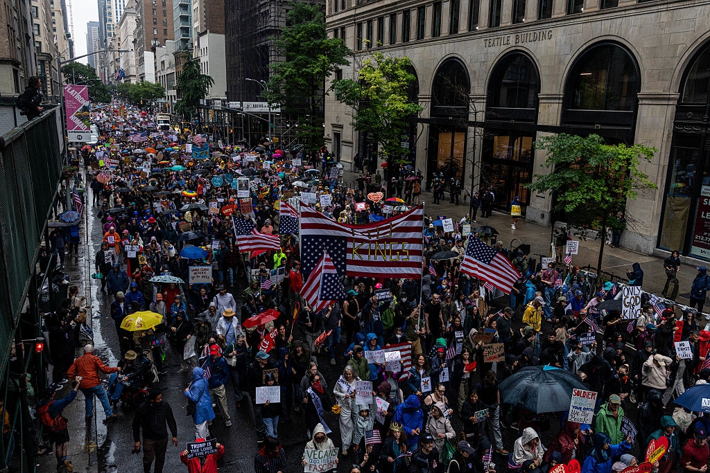 Crowd marching in the rain on city street, holding protest signs and American flags, with large buildings in the background