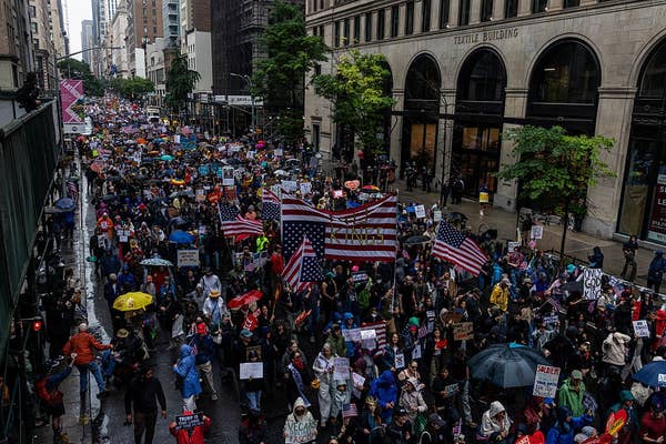 Crowd marching in the rain on city street, holding protest signs and American flags, with large buildings in the background