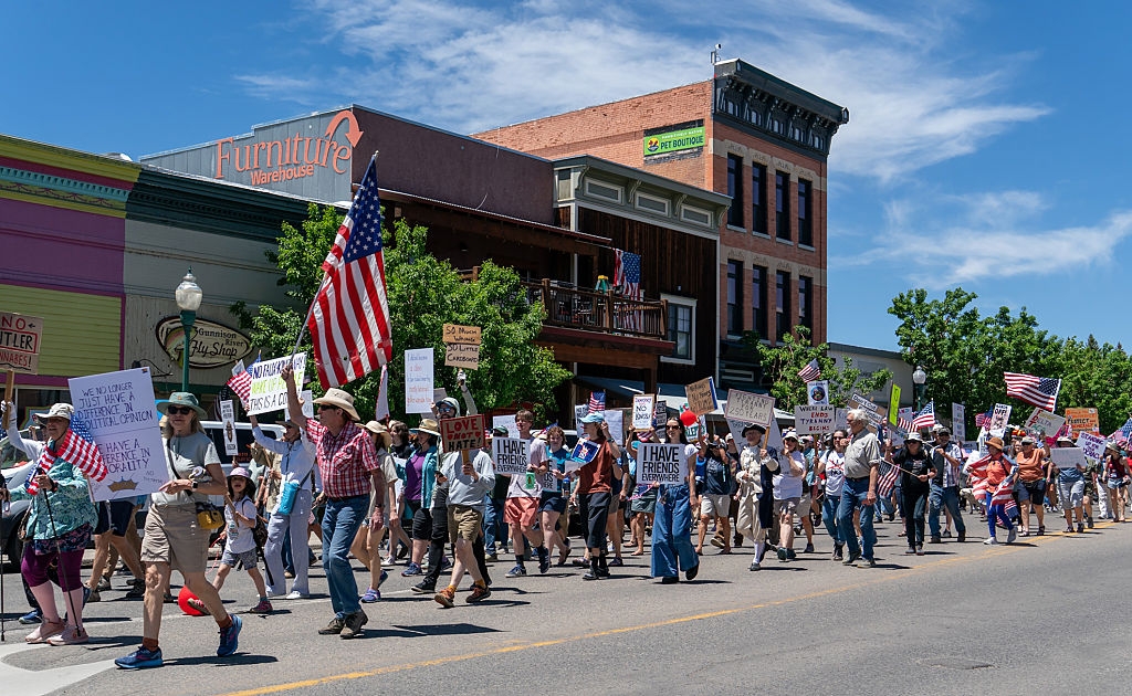 Large crowd marches down a street holding signs and American flags during a protest, with a furniture store visible in the background