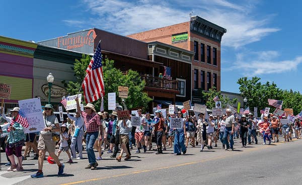 Large crowd marches down a street holding signs and American flags during a protest, with a furniture store visible in the background