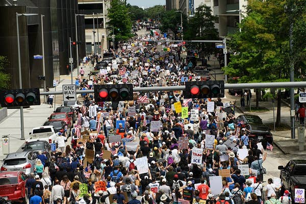 Large crowd of protesters fill a city street, holding signs and banners, participating in a march for social or political change