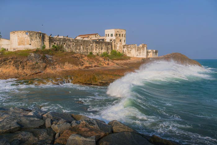 Waves hit rocks near Elmina Castle, a historic coastal fortress, with the building's weathered walls overlooking the ocean in the background