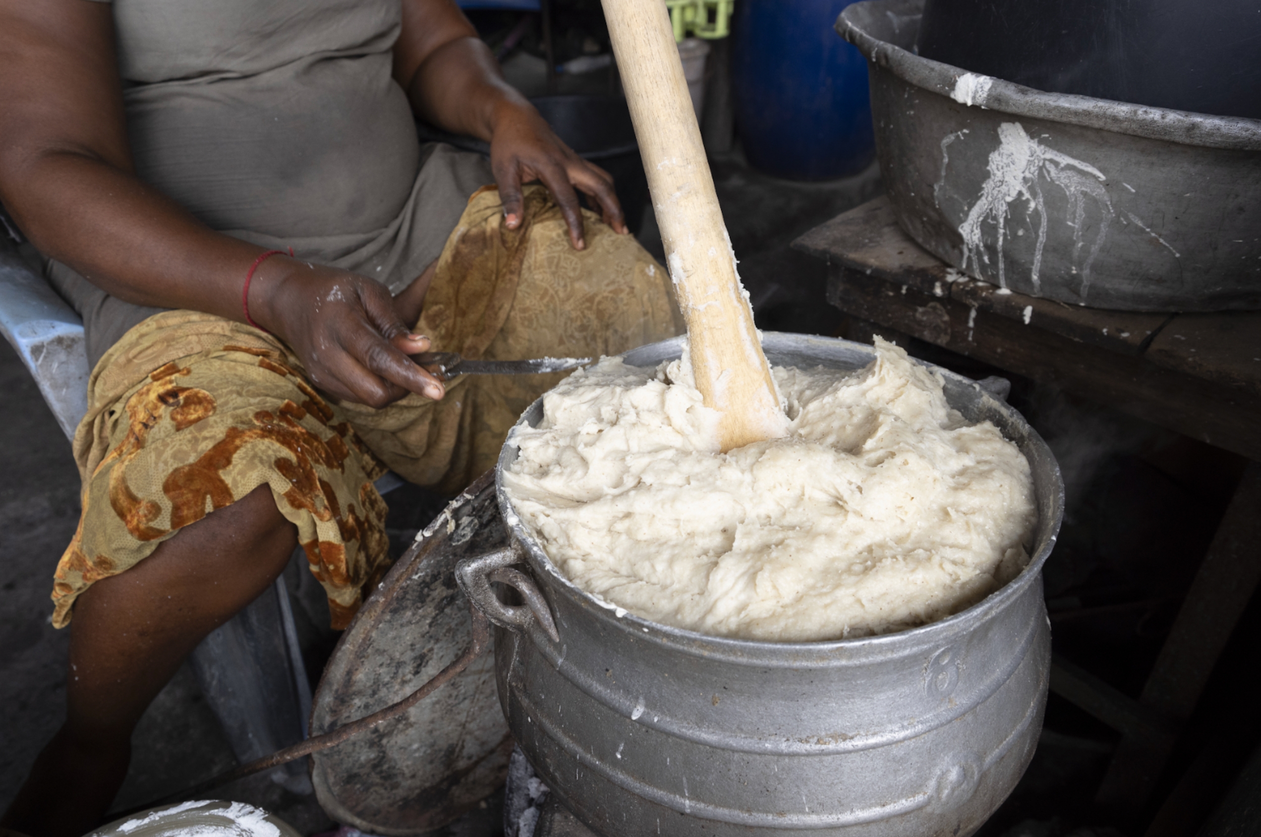 Person stirring a large pot of traditional food dough with a wooden paddle, sitting at a roadside setup