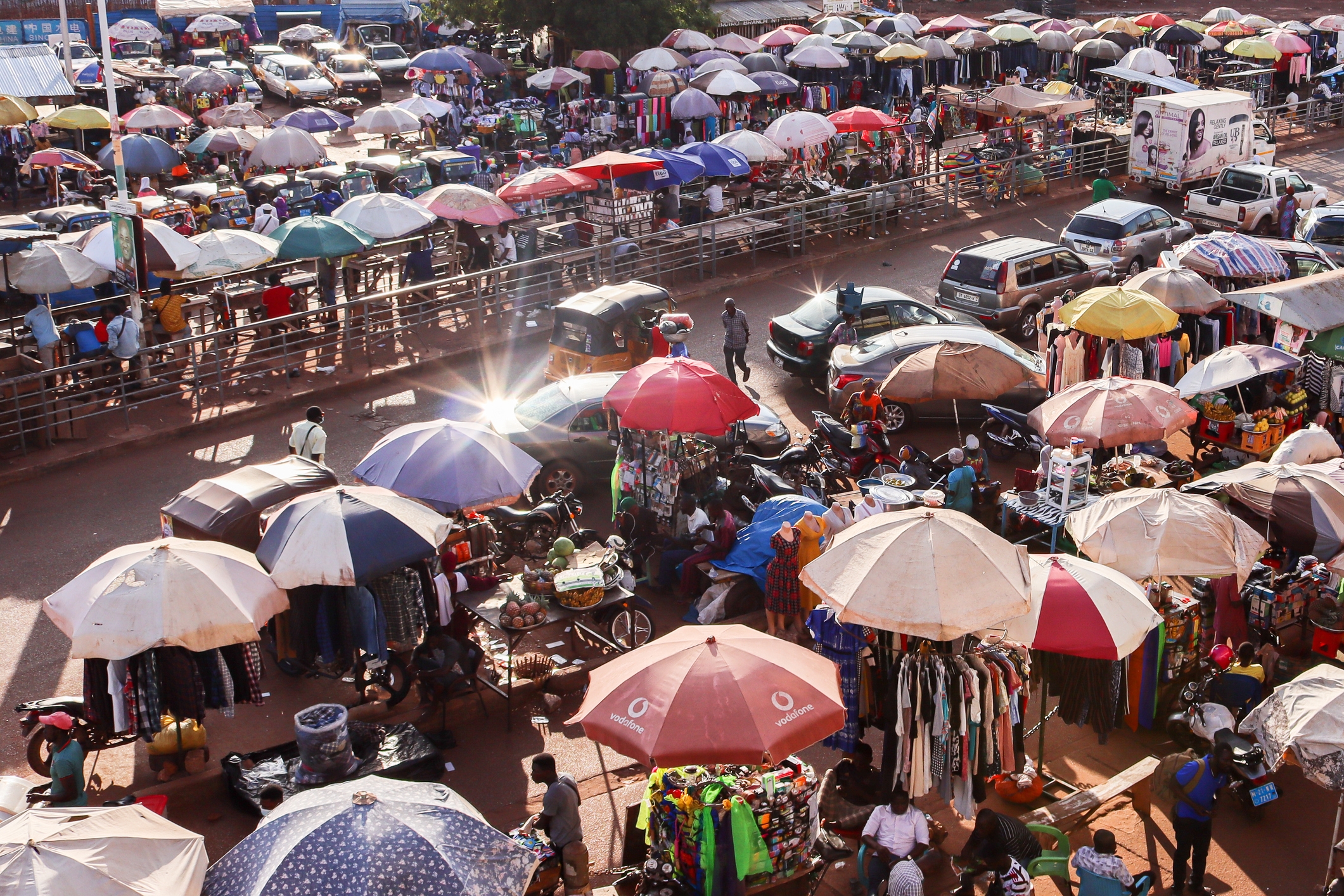 Bustling outdoor market with various stalls under umbrellas, selling clothes and goods. People shop and walk among parked cars