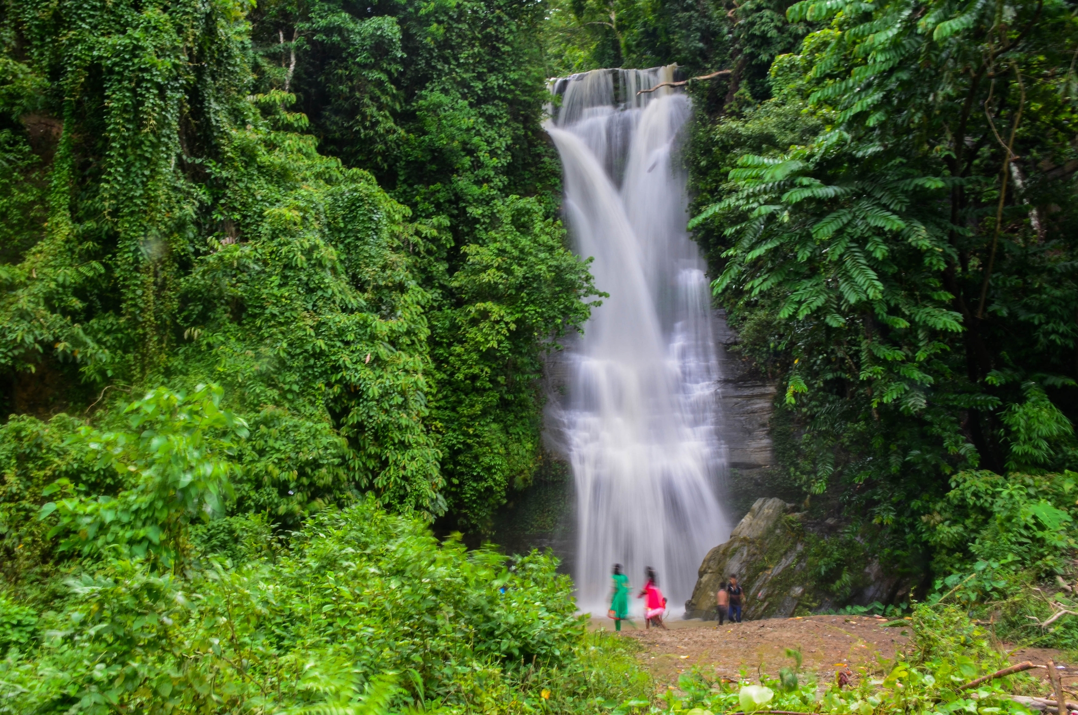Waterfall cascading down rocks surrounded by lush greenery, with four people standing at the base, capturing the serene and natural beauty of the scene