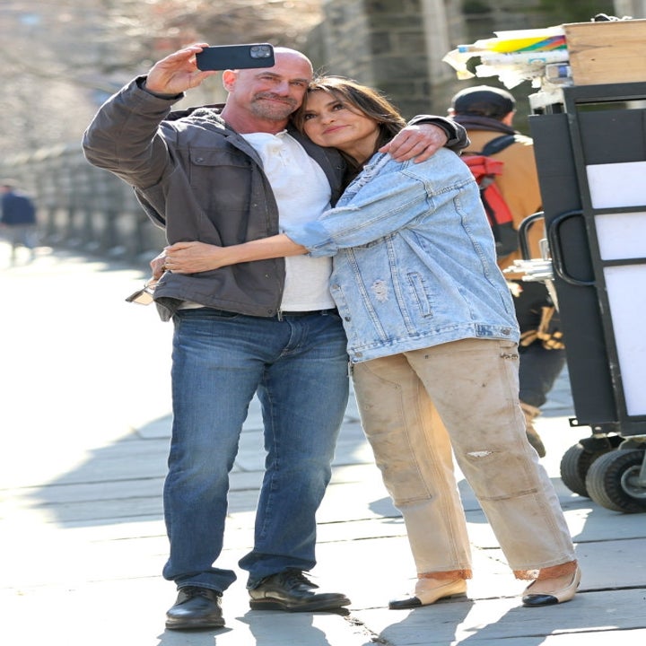 Two people taking a selfie. The man wears a jacket and jeans, while the woman wears a denim jacket and beige pants. They are embracing outdoors