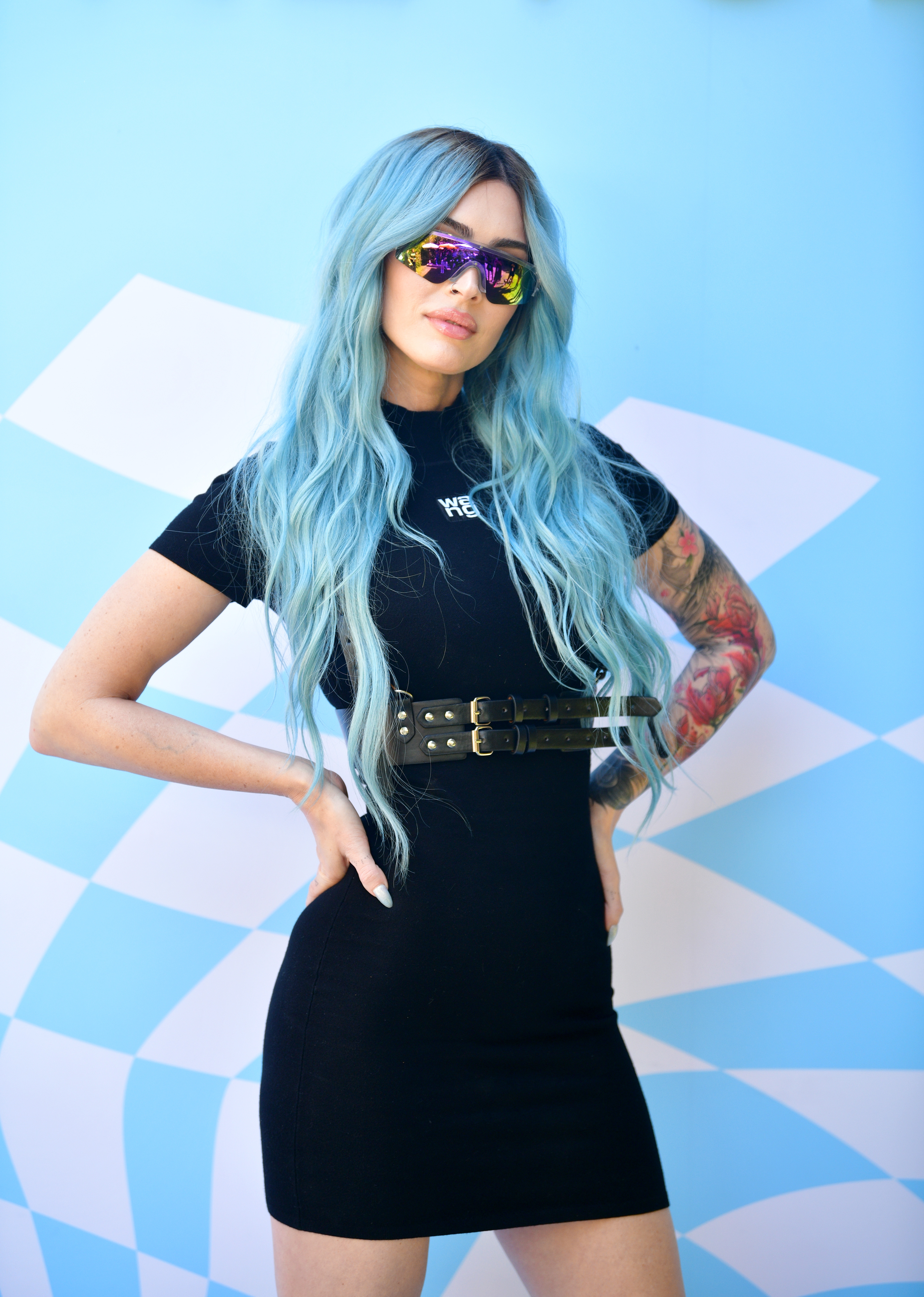 Megan Fox with long, blue hair wearing a black dress and sunglasses stands confidently with hands on hips against a patterned background