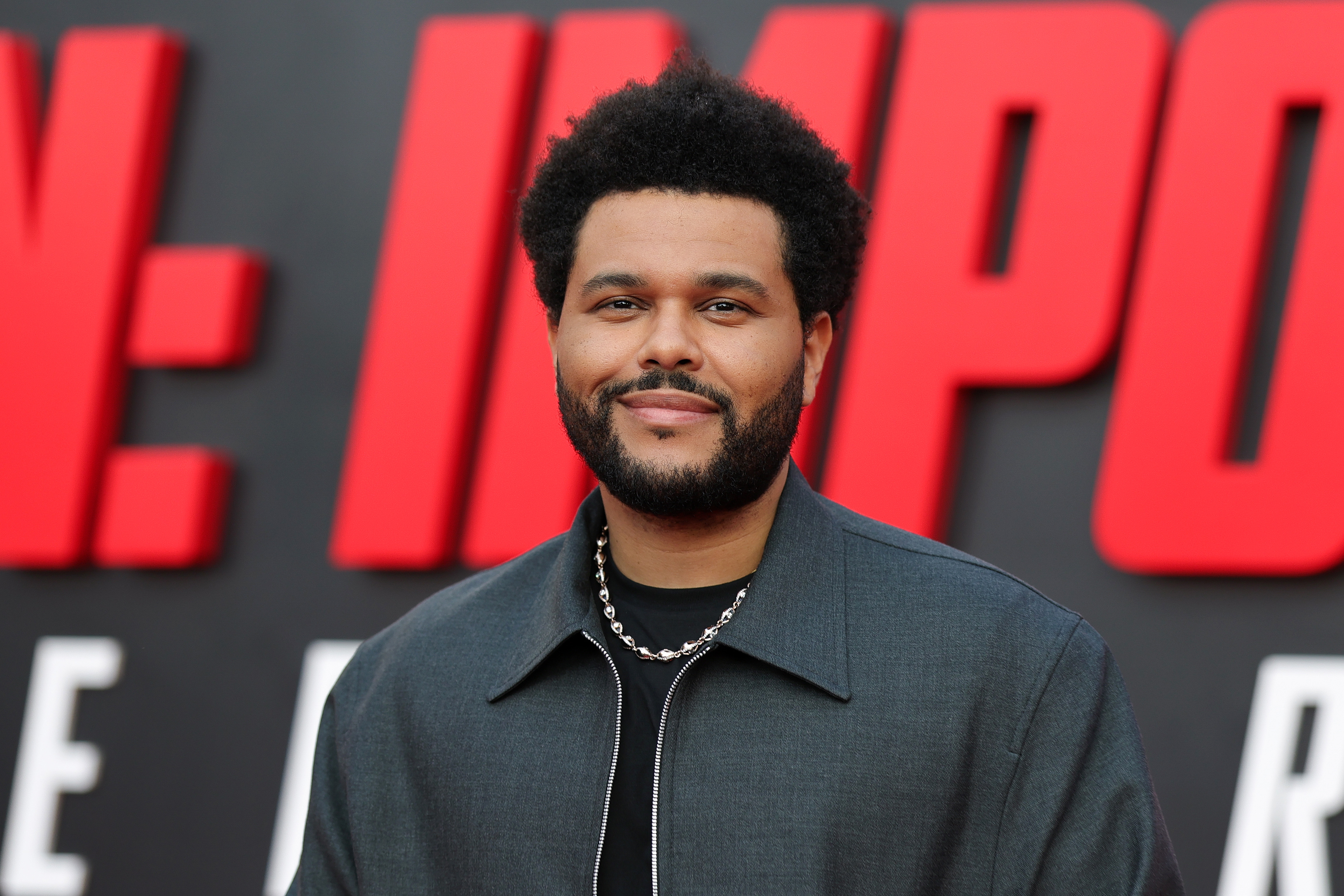 The Weeknd smiling on a red carpet, wearing a sleek jacket and a chain necklace against a backdrop with large, bold lettering