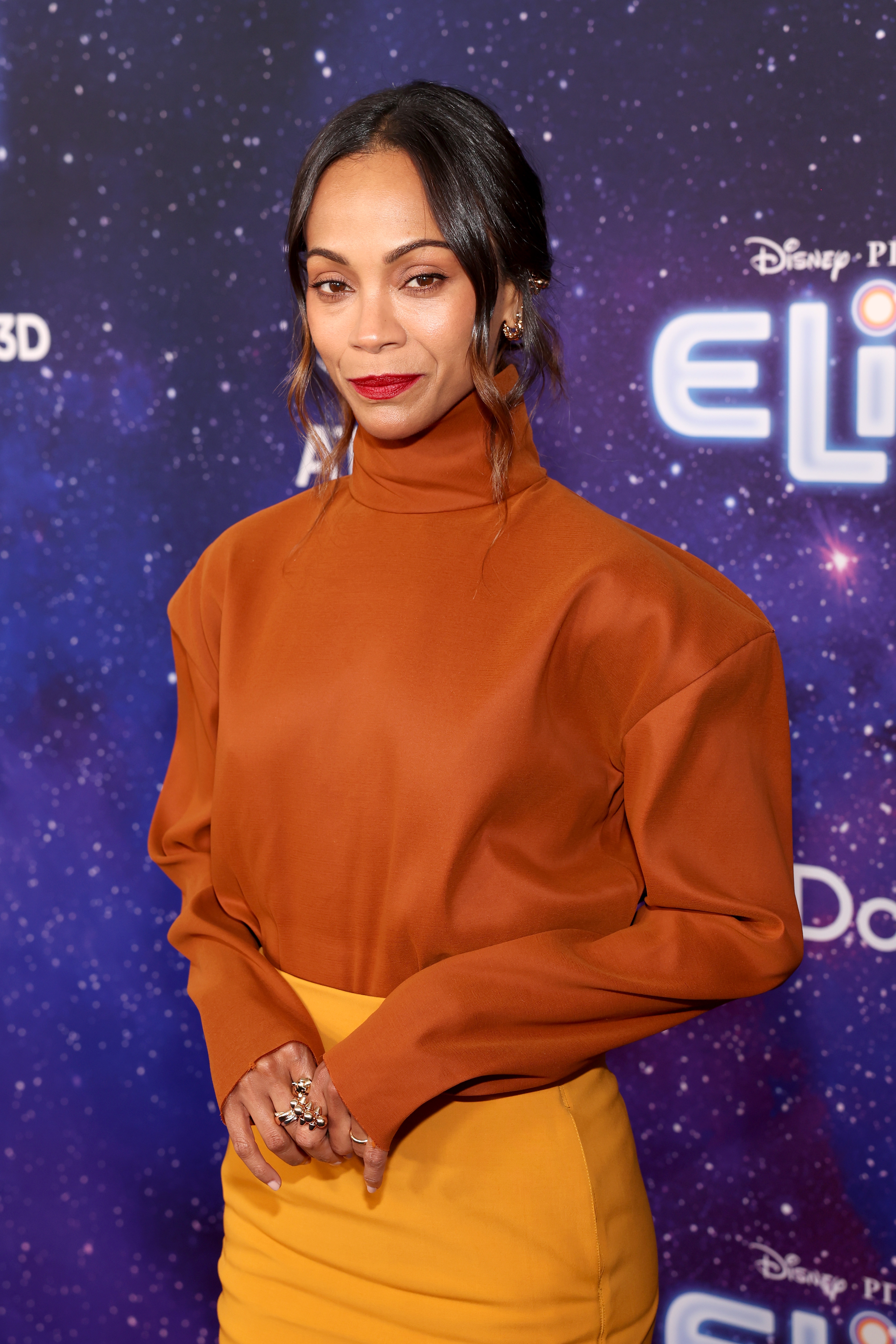 Zoe Saldaña posing on a red carpet in a high-neck top and skirt, with a cosmic-themed backdrop in the background
