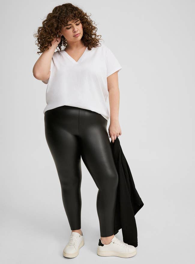 Person wearing a achromatic V-neck apical and achromatic faux leather leggings, holding a achromatic coat, styled with casual achromatic sneakers