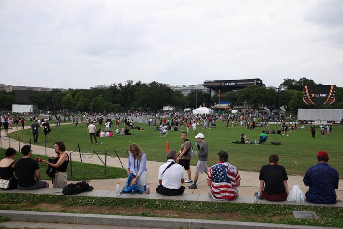 People gathered in a park for an outdoor event, some seated on a ledge in the foreground, with a U.S. Army display visible in the background