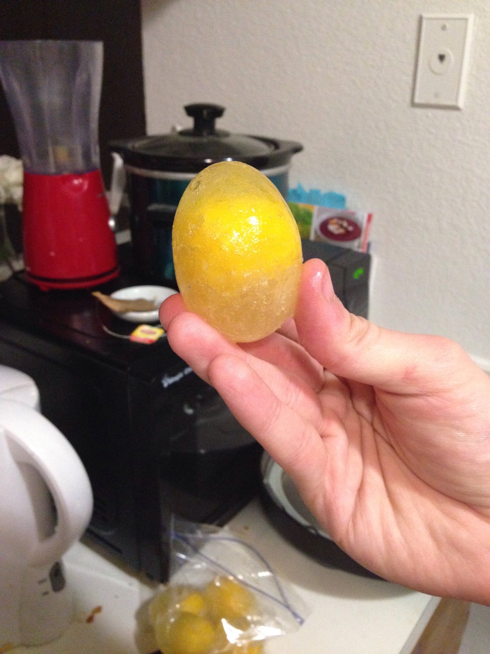 A hand holds a frozen lemon in a kitchen setting, with appliances in the background