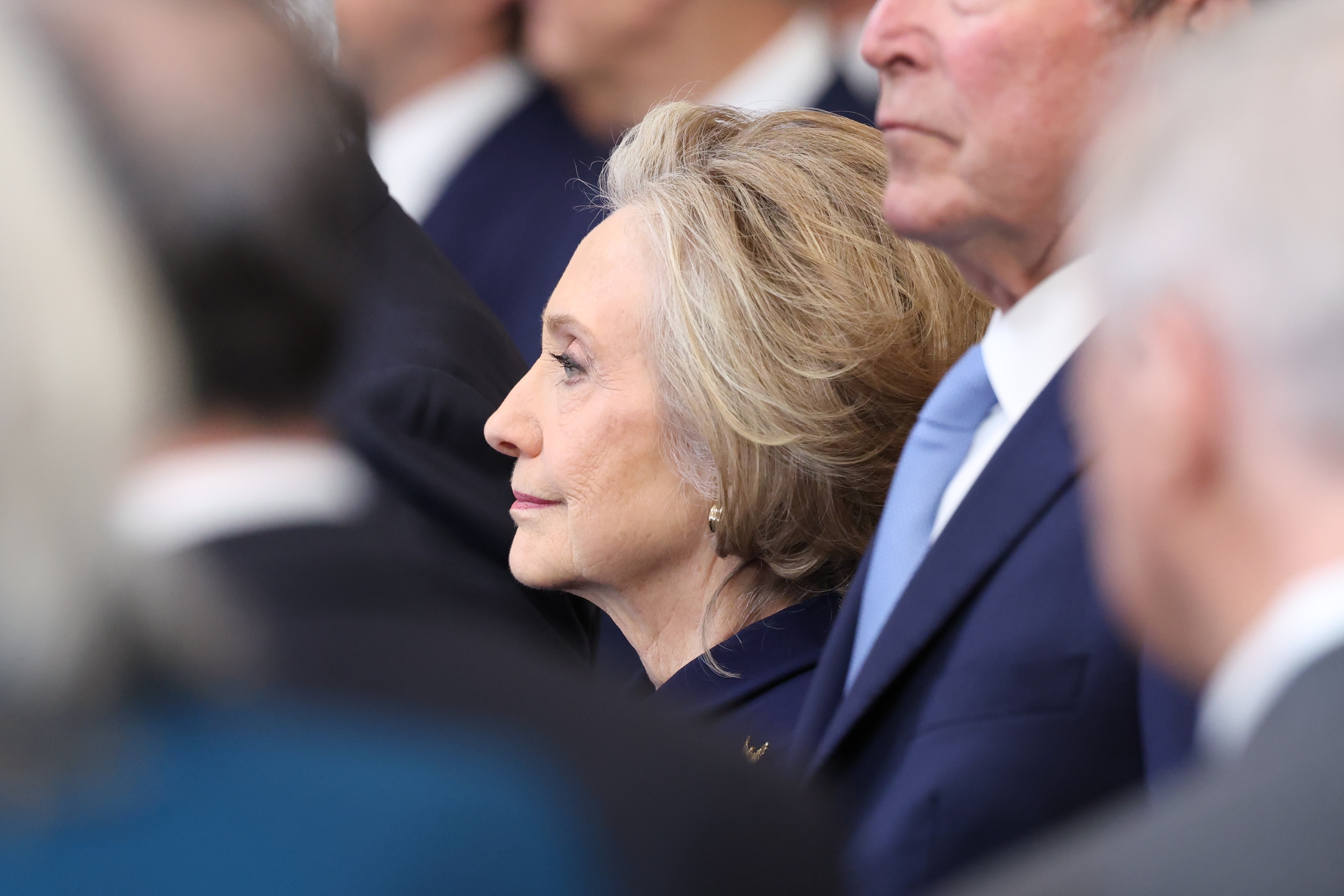 Person in profile, dressed formally, attending a solemn event. Others in similar attire are partially visible in the background