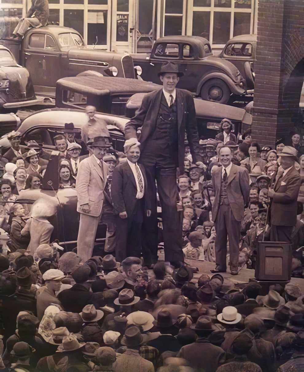 A historical photo of a tall man standing among a crowd, surrounded by onlookers in vintage attire, with classic cars in the background