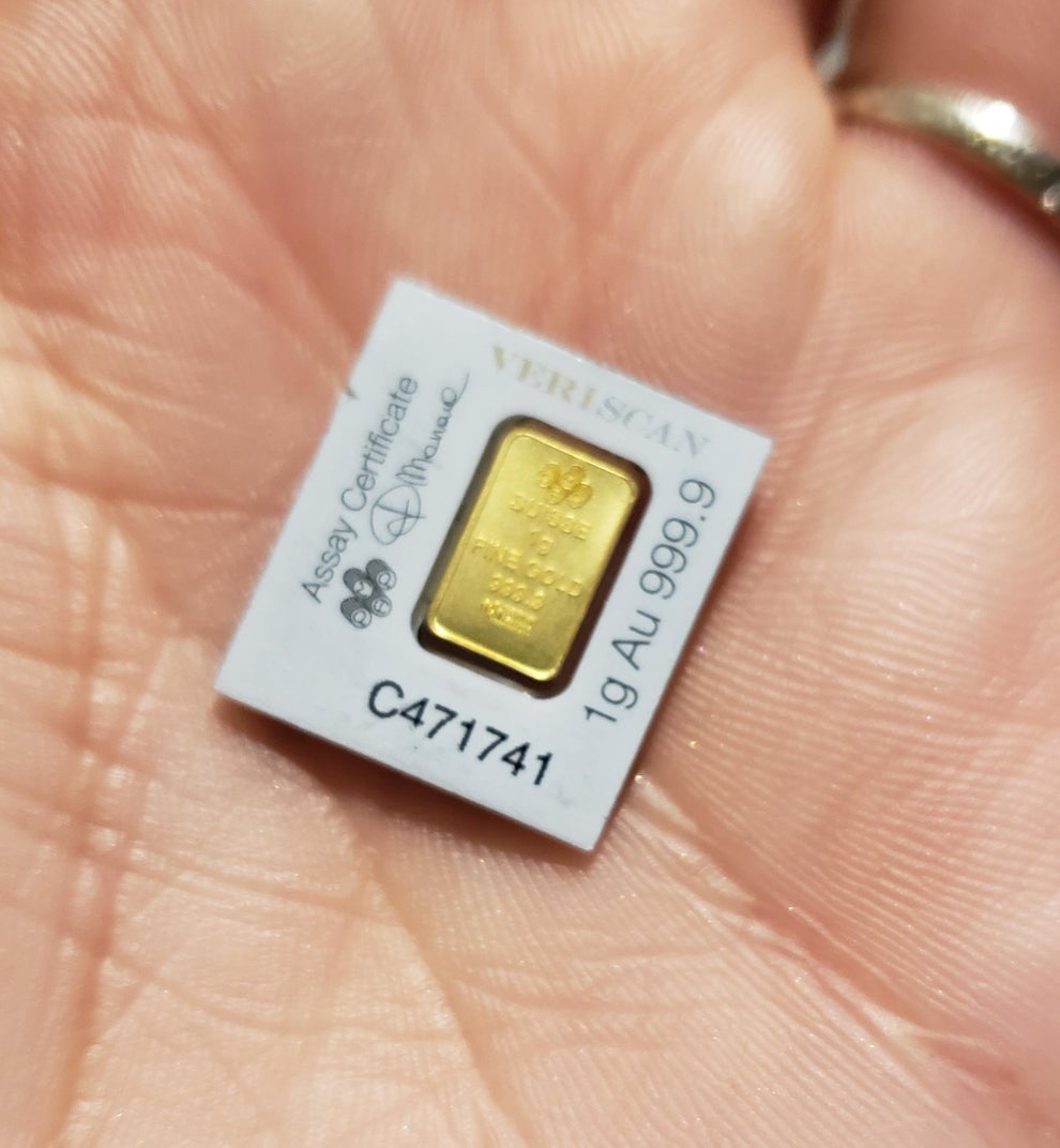 A hand holds a small gold bar with a certificate indicating 1g of 999.9 pure gold, embossed numbers, and text