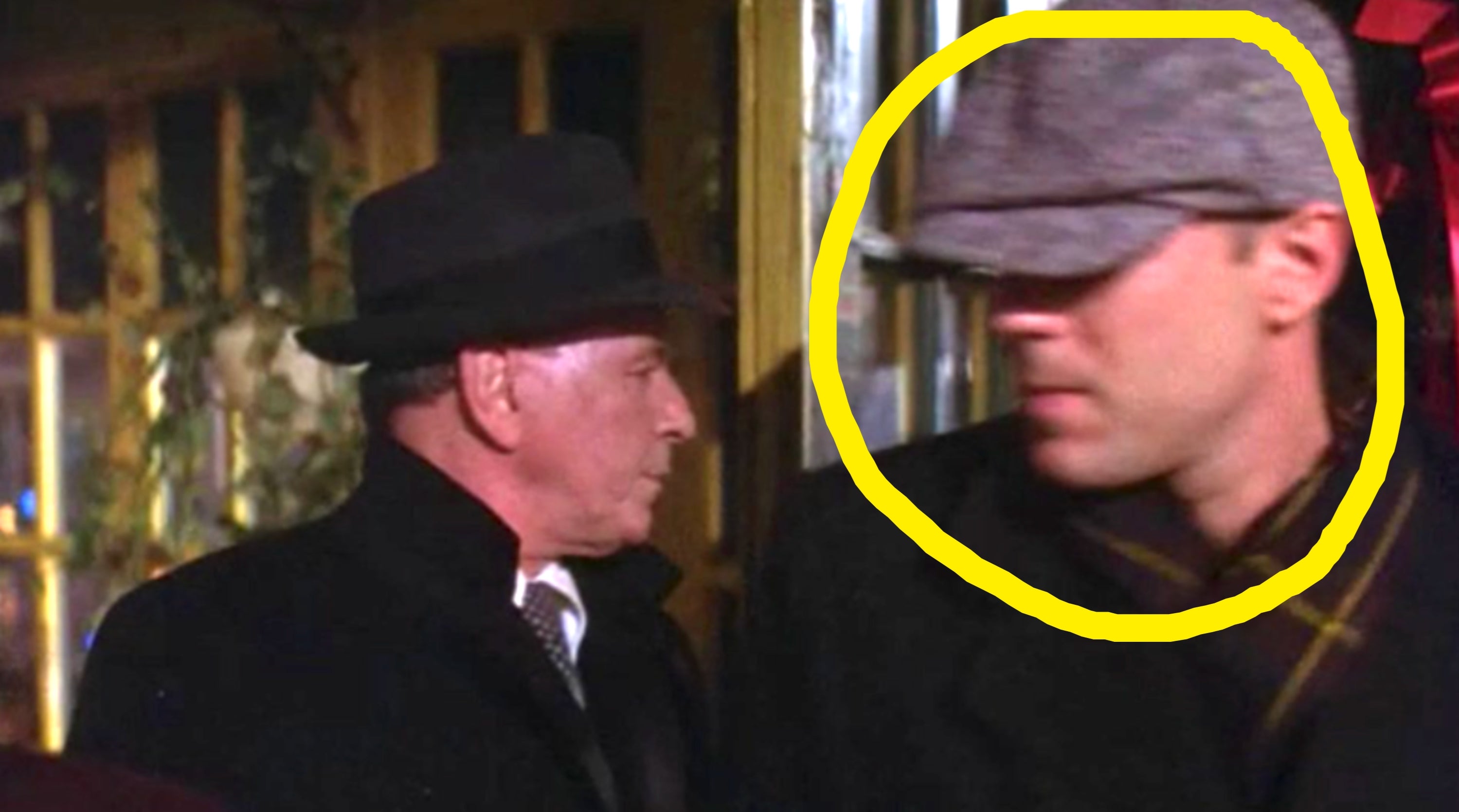Two people wearing coats and hats stand outside a building. One looks to the side while the other looks forward
